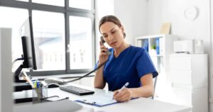 Healthcare professional talking on the phone while taking notes in a medical office, answering questions and providing guidance about early pregnancy symptoms and pregnancy testing.